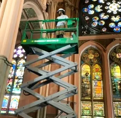 A man is on a scissor lift in front of stained glass windows.